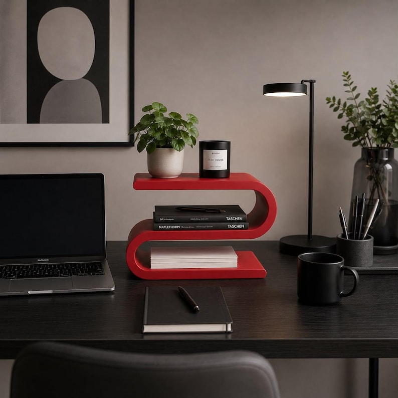 May include: A bright red S-shaped wooden shelf with three horizontal tiers. The top tier holds a small green potted plant and a black candle. The middle tier contains two stacked black books. The bottom tier holds a stack of white paper.