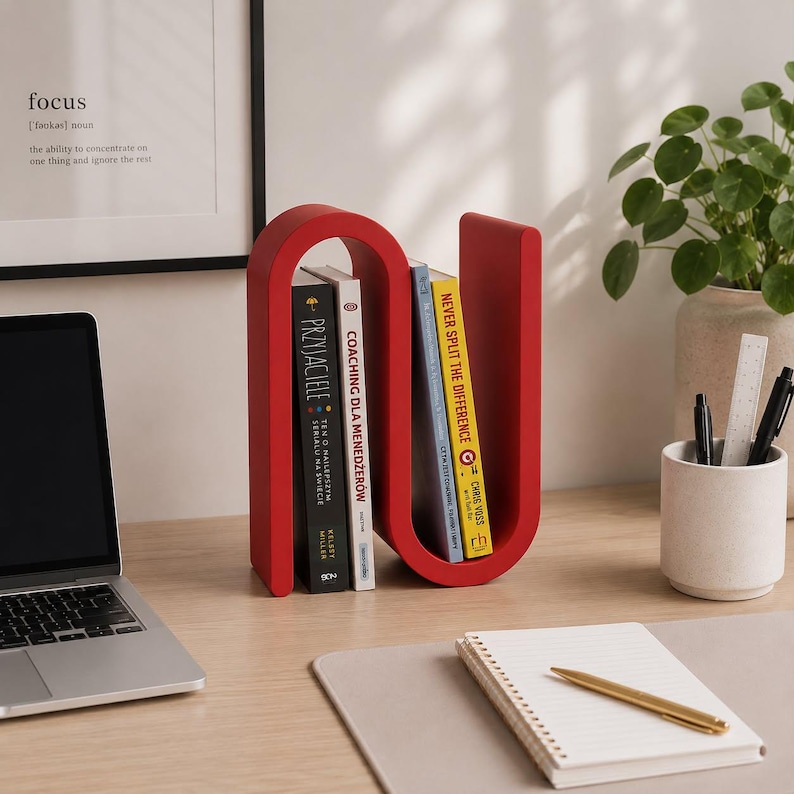 May include: A bright red, S-shaped metal bookend holding four books upright. The bookend has a smooth, curved design that creates two distinct sections for organizing reading materials on a desk surface alongside a laptop and stationery.