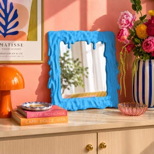 Op de afbeelding: Een rechthoekige spiegel met een felblauw frame met een getextureerd, druipend vloeistofontwerp. De spiegel staat rechtop, reflecteert een wazige binnenruimte met groen en is omringd door diverse decoratieve objecten voor in huis.