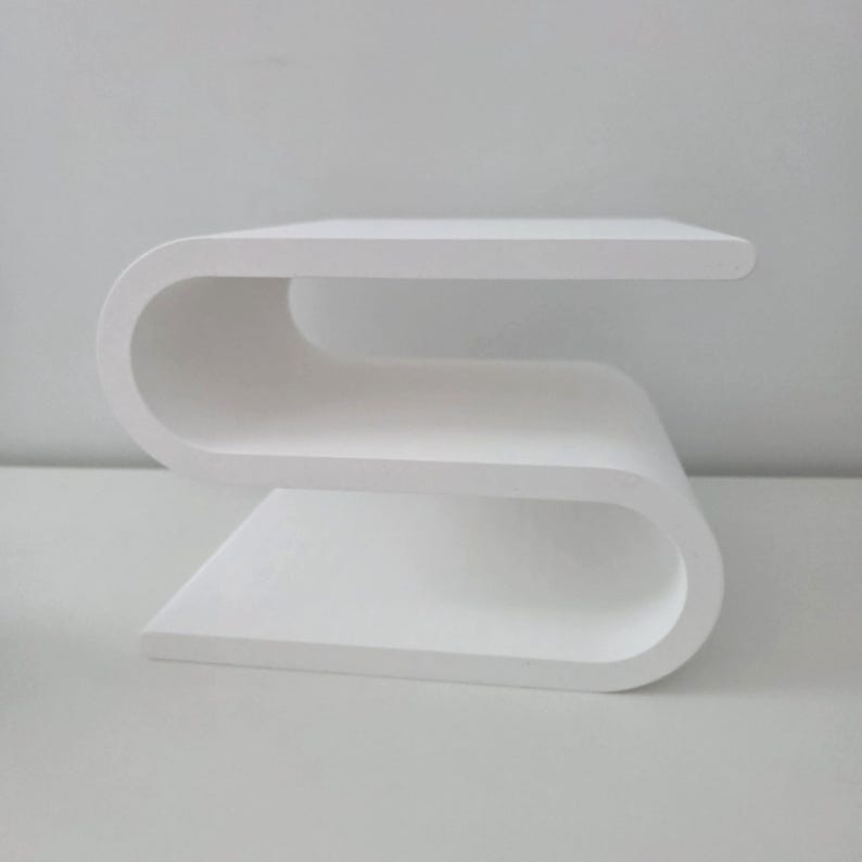 May include: A white side table featuring a continuous S-shaped design with a flat top surface, a middle shelf, and a flat base. The structure is formed from a single piece of material with smooth, rounded curves connecting each horizontal level.