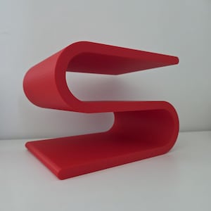 May include: A bright red S-shaped shelf featuring a continuous, curved design with two flat horizontal surfaces. The structure is formed from a single piece of material, creating a smooth, flowing silhouette that provides two distinct levels.