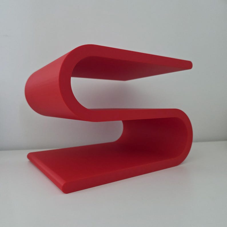 May include: A bright red S-shaped shelf featuring a continuous, curved design with two flat horizontal surfaces. The structure is formed from a single piece of material, creating a smooth, flowing silhouette that provides two distinct levels.