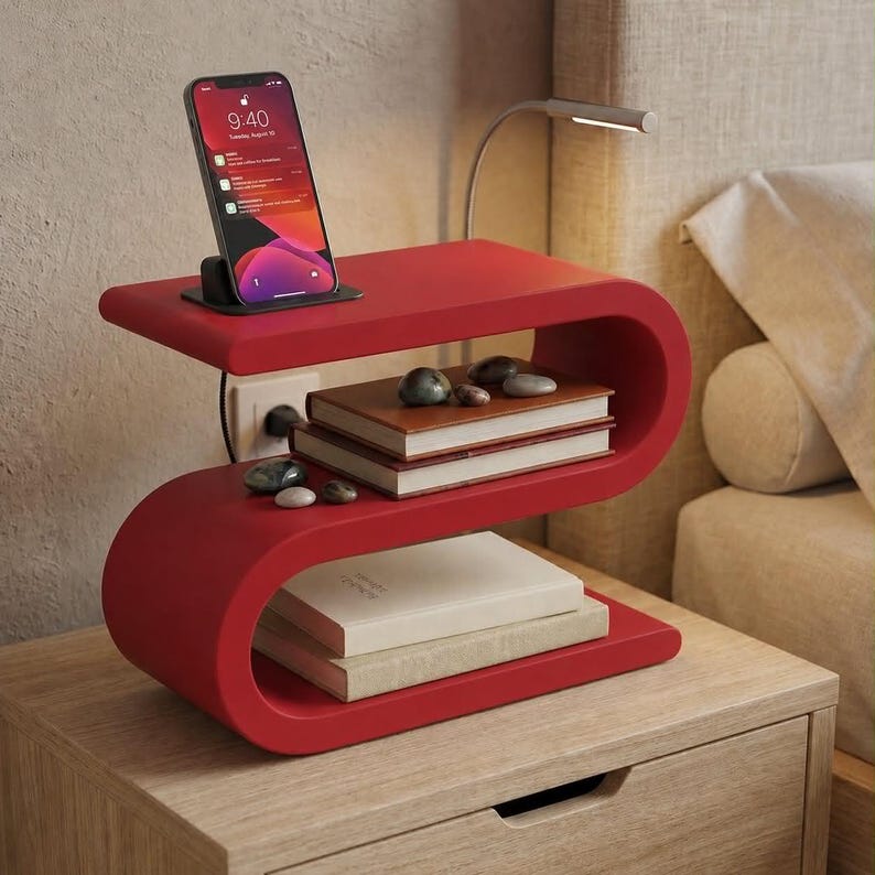 May include: A red S-shaped bedside table featuring a top surface with a smartphone in a charging dock, a middle shelf holding two books and small stones, and a bottom shelf with two additional books. The table has a smooth, matte finish.