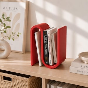 May include: A bright red, S-shaped book rack made of a smooth, solid material. The rack holds three books upright, including titles titled Przyjaciele and Coaching dla Menedżer&oacute;w, with the curved structure providing support on both sides.