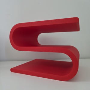 May include: A bright red S-shaped shelf with a smooth, matte finish. The structure features two horizontal surfaces connected by a continuous, curved frame, creating an open design that allows for storage or display on both the top and middle levels.