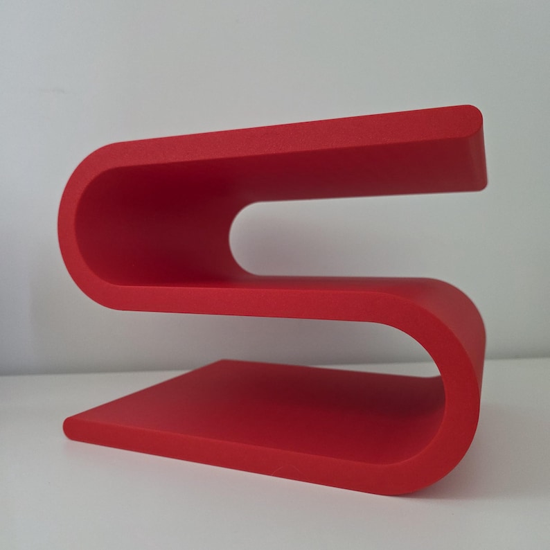 May include: A bright red S-shaped shelf with a smooth, matte finish. The structure features two horizontal surfaces connected by a continuous, curved frame, creating an open design that allows for storage or display on both the top and middle levels.