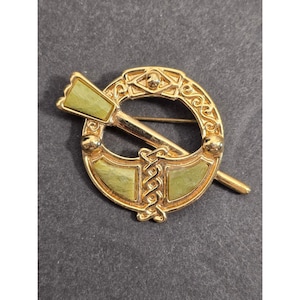 May include: A gold-toned brooch with a circular design, featuring a central pin and two green rectangular accents. The brooch has intricate detailing and a decorative pin, suitable for adding a touch of elegance to any outfit.