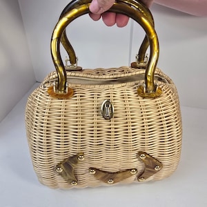 May include: A light tan woven wicker handbag featuring two rounded amber-colored tortoiseshell handles. The front is decorated with three chevron-shaped tortoiseshell accents secured by gold-tone metal studs and a central gold-tone metal clasp.