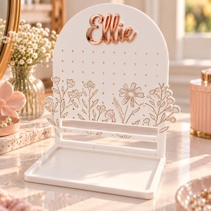 May include: A white arched jewelry display stand featuring a rose gold metallic nameplate that reads Ellie. The stand has a perforated upper section for earrings, a floral line art design, and a rectangular base tray for small accessories.