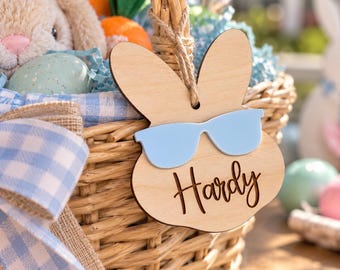 May include: A light-colored wooden tag shaped like a rabbit head with long ears, wearing light blue sunglasses. The name Hardy is engraved in dark brown script on the tag, which hangs from a woven wicker basket with a blue gingham ribbon.