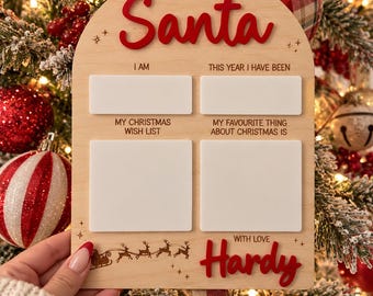 May include: An arched wooden sign with the text Dear Santa in red, featuring four blank white rectangular spaces for writing. Below, the text reads My Christmas Wish List and My Favourite Thing About Christmas Is, with the name Hardy in red.