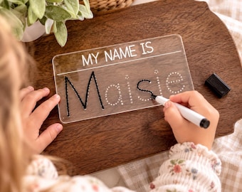 Pode incluir: Uma placa de prática retangular de acrílico transparente com o texto impresso em branco MY NAME IS e o nome Maisie em pontilhado. Um marcador apagável preto está sendo usado para traçar as letras na superfície da placa.