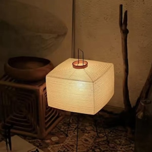 May include: A square paper lantern emitting a warm light. It features a brown metal handle and top, supported by black metal legs. The lamp is a decorative lighting fixture, ideal for creating a cosy atmosphere. The lamp's dimensions are approximately 30 cm x 30 cm.