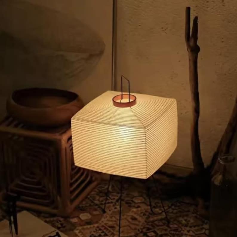 May include: A square paper lantern emitting a warm light. It features a brown metal handle and top, supported by black metal legs. The lamp is a decorative lighting fixture, ideal for creating a cosy atmosphere. The lamp's dimensions are approximately 30 cm x 30 cm.