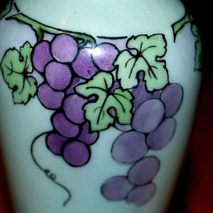 May include: A white ceramic vase with a hand-painted design of purple grapes and green leaves.
