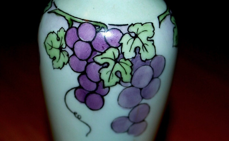 May include: A white ceramic vase with a hand-painted design of purple grapes and green leaves.