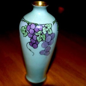 May include: A small, light blue ceramic vase with a gold rim. The vase is decorated with a painted design of purple grapes and green leaves.