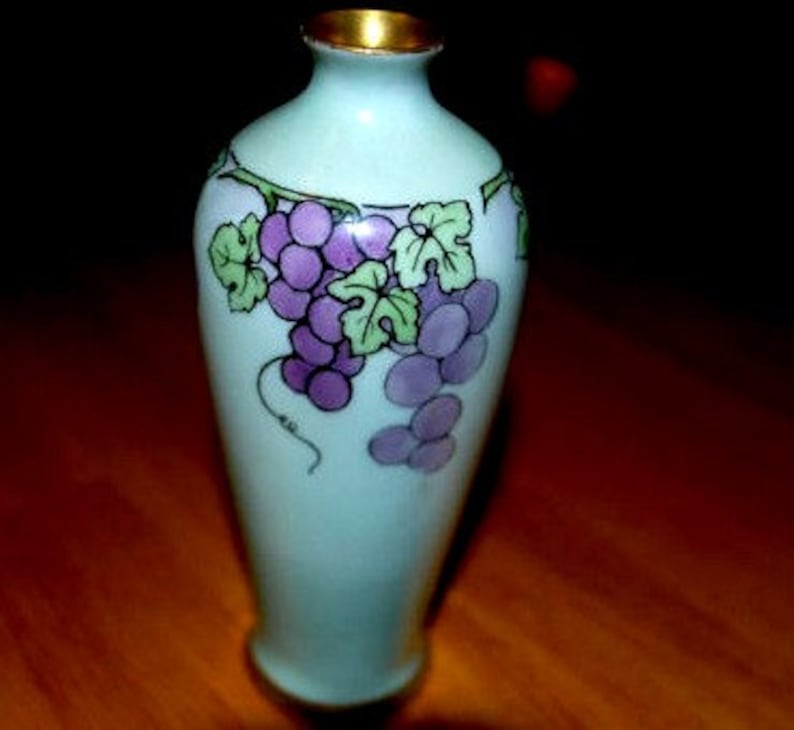 May include: A small, light blue ceramic vase with a gold rim. The vase is decorated with a painted design of purple grapes and green leaves.