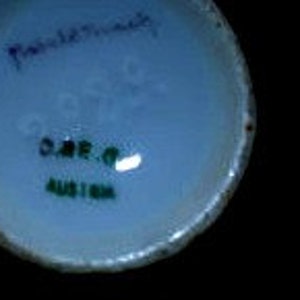 May include: Close-up of the base of a white ceramic mug with green and blue lettering that reads "Handpainted" and "O.A.R.O. Austria".