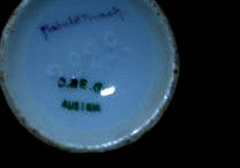 May include: Close-up of the base of a white ceramic mug with green and blue lettering that reads "Handpainted" and "O.A.R.O. Austria".
