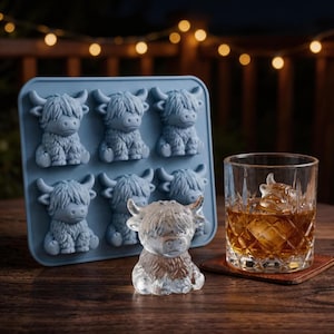 Puede incluir: Una bandeja de cubitos de hielo de silicona azul con seis moldes en forma de vaca de las Tierras Altas. Un cubito de hielo transparente en forma de vaca de las Tierras Altas se encuentra junto a un vaso de líquido ámbar con un cubito de hielo en forma de vaca. El vaso está sobre un posavasos marrón.