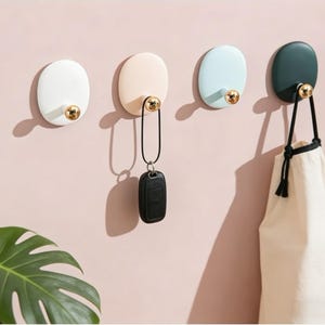 Puede incluir: Cuatro ganchos de pared ovalados en blanco, melocotón, azul claro y verde oscuro, cada uno con un pomo dorado. Una llave de coche negra y una bolsa crema cuelgan de dos de los ganchos. Los ganchos están montados en una pared rosa.