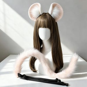 May include: A set of faux fur animal ears on a headband and a long, matching faux fur tail with a black adjustable strap and plastic buckle. The ears are pale pink with white centres, and the tail is a soft, pale pink colour throughout.