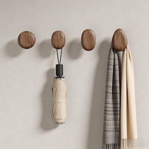 Könnte beinhalten: Vier hölzerne Wandhaken in einer Reihe. Die Haken sind dunkelbraun und haben eine glatte, abgerundete Form. Ein Regenschirm und zwei Schals hängen an den Haken. Der Regenschirm ist beige und schwarz, und die Schals sind grau und cremefarben.