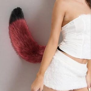 Op de afbeelding: Een rood en zwarte nepbont staart, bevestigd aan een grijze muur. De staart is gebogen en pluizig, met een zwarte punt en een verloop naar een diep rood. Dit is een kostuumaccessoire.