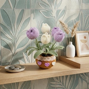 May include: A crochet tulip arrangement featuring two light purple and two cream-coloured flowers with green leaves. The flowers are housed in a brown pot decorated with a white lace trim and small purple and white heart-shaped accents.