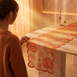 May include: A white table runner featuring an orange embroidered pattern of pumpkins, leaves, and swirls. The runner has a scalloped edge finished with orange trim, draped over a white marble table surface in a warm, softly lit indoor setting.