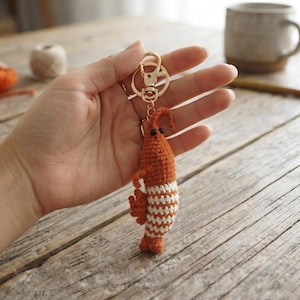 May include: A crochet shrimp keychain featuring a gold-toned metal lobster clasp. The shrimp is crafted with orange and white yarn in a striped pattern, with small black beads for eyes and a textured, segmented body design hanging from the ring.