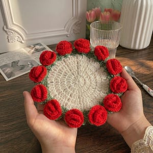 Puede incluir: Un posavasos redondo de ganchillo color crema con un borde de once pequeñas rosas rojas tridimensionales. El centro del posavasos está tejido con un patrón circular y las rosas están unidas a una base de ganchillo color verde salvia.