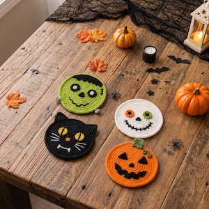 Puede incluir: Cuatro posavasos redondos de ganchillo con diseños de Halloween: un monstruo de Frankenstein verde, un gato negro con ojos amarillos, una calavera blanca con ojos desiguales y una calabaza naranja con tallo verde, dispuestos sobre una superficie de madera.