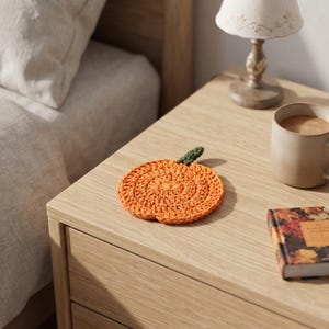May include: A round, orange, crocheted coaster shaped like a pumpkin, featuring a small, dark green crocheted stem at the top. The coaster is crafted with a textured, circular stitch pattern, creating a flat, functional piece for resting drinks.
