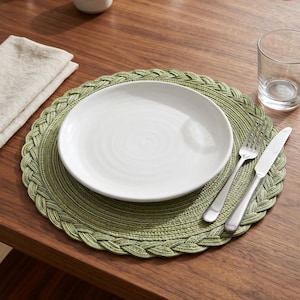 May include: A round sage green placemat featuring a braided edge design. The placemat is topped with a plain white ceramic dinner plate, a stainless steel fork, and a stainless steel knife, creating a complete table setting arrangement.