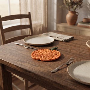 May include: A round, orange, crocheted pumpkin-shaped coaster with a small green stem detail. The coaster is placed on a dark brown wooden dining table between two speckled ceramic plates and two silver metal forks, with a napkin nearby.