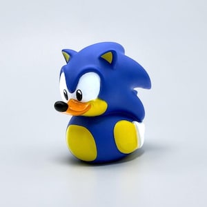 May include: A blue rubber duck toy designed to resemble a hedgehog character. The figure features a bright blue body, a yellow belly, a yellow muzzle, a small black beak, and large white eyes with black pupils, all set against a plain background.