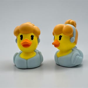 May include: Two yellow rubber duck toys with orange beaks and brown hair. The ducks wear light blue outfits and have black eyes. One duck has its hair down, and the other has its hair up in a bun.