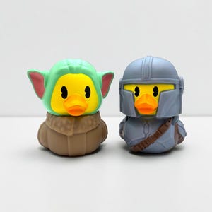 Op de afbeelding: Twee rubberen eendjes ontworpen als personages. Eén eendje heeft een lichtgroene capuchon met grote oren en een bruine jas. Het andere eendje draagt een grijze helm en bijpassend grijs harnas met een bruine riem over de borst.