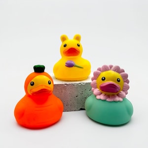 May include: Three rubber duck toys in bright colors. One is orange with a green hat, another is yellow with a purple flower, and the third is yellow with a pink flower and green base. The ducks are on a small, gray block against a white background.