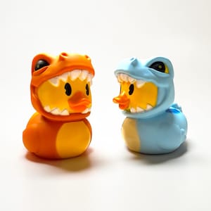 May include: Two rubber duck toys, one orange and one blue, each wearing a dinosaur costume. The orange duck has an orange dinosaur head with white teeth, while the blue duck has a blue dinosaur head with white teeth. Both ducks have yellow bodies and orange beaks.