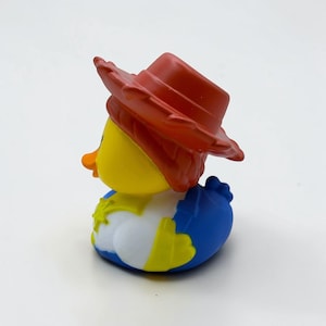 K&ouml;nnte beinhalten: Ein gelbes Quietscheentchen mit einem roten Cowboyhut, einem wei&szlig;en Hemd mit einem gelben Sheriffstern und einer blauen Hose. Die Ente hat einen orangefarbenen Schnabel und eine glatte, geformte Oberfl&auml;che, die an eine Figur erinnert.