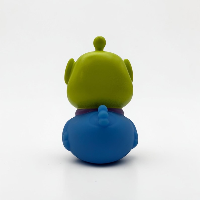 K&ouml;nnte beinhalten: Eine kleine, glatte, drei&auml;ugige Alien-Figur von hinten betrachtet. Die Figur hat einen lindgr&uuml;nen Kopf mit einer kleinen Antenne oben, einen violetten Kragen und einen einfarbigen blauen K&ouml;rper. Sie zeigt abgerundete Ohren und ein schwanzartiges Detail.
