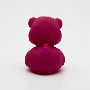 K&ouml;nnte beinhalten: Eine massive, leuchtend magentafarbene Gummiente-Figur, von hinten betrachtet. Das Objekt zeigt einen abgerundeten Kopf mit zwei ausgepr&auml;gten Ohren, einen glatten, geschwungenen K&ouml;rper und ein kleines, erhabenes Schwanzdetail in der Mitte des unteren R&uuml;ckens.