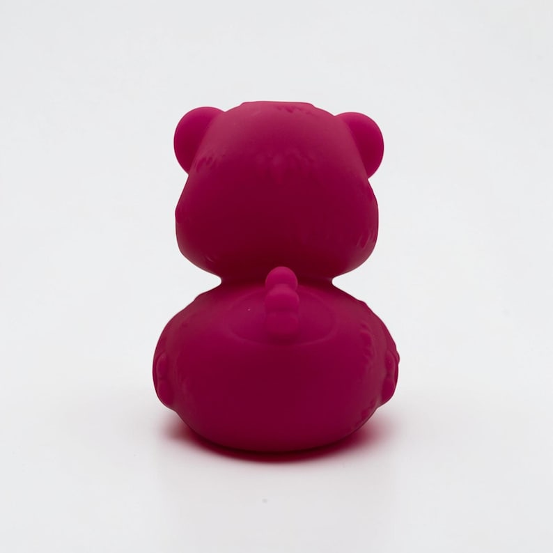K&ouml;nnte beinhalten: Eine massive, leuchtend magentafarbene Gummiente-Figur, von hinten betrachtet. Das Objekt zeigt einen abgerundeten Kopf mit zwei ausgepr&auml;gten Ohren, einen glatten, geschwungenen K&ouml;rper und ein kleines, erhabenes Schwanzdetail in der Mitte des unteren R&uuml;ckens.