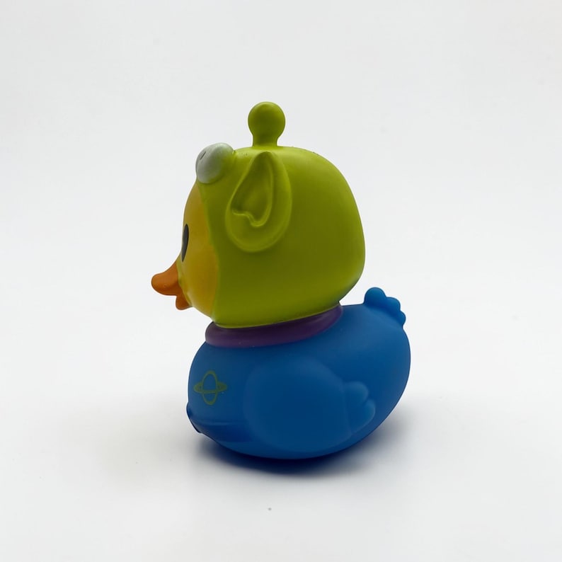 K&ouml;nnte beinhalten: Ein Quietscheentchen mit einer lindgr&uuml;nen Alien-Kapuze mit kleiner Antenne, einem leuchtend orangefarbenen Schnabel und einem blauen K&ouml;rper. Der blaue Bereich ist mit einem violetten Kragen und einem kleinen gelben Planeten-Symbol an der Seite versehen.