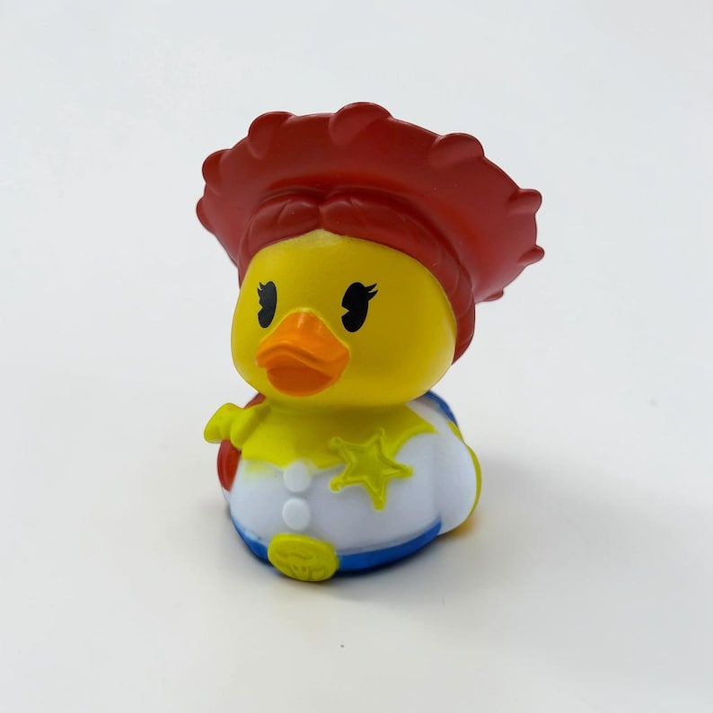 K&ouml;nnte beinhalten: Ein gelbes Quietscheentchen im Cowgirl-Stil. Die Ente tr&auml;gt einen gro&szlig;en roten Hut, ein wei&szlig;es Hemd mit einem gelben Sheriffstern-Abzeichen und eine blaue Basis. Sie hat schwarze Wimpern, einen orangefarbenen Schnabel und wei&szlig;e Kn&ouml;pfe am Hemd.