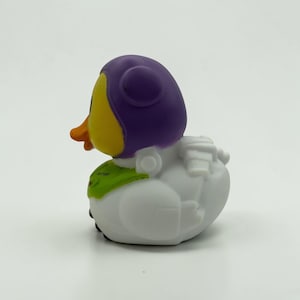 K&ouml;nnte beinhalten: Ein Quietscheentchen mit violetter Kapuze, gelbem Gesicht und orangefarbenem Schnabel. Der K&ouml;rper ist wei&szlig; mit einem gr&uuml;nen Brustteil, auf dem der Text LIGHTYEAR steht. Die Figur hat geformte wei&szlig;e Fl&uuml;gel und ein Rucksackdetail am R&uuml;cken.