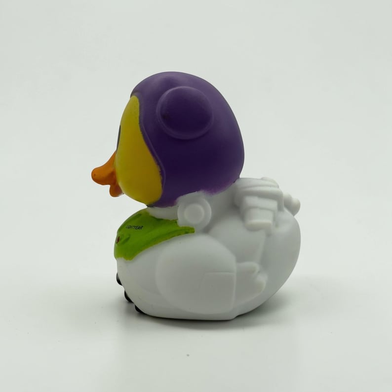 K&ouml;nnte beinhalten: Ein Quietscheentchen mit violetter Kapuze, gelbem Gesicht und orangefarbenem Schnabel. Der K&ouml;rper ist wei&szlig; mit einem gr&uuml;nen Brustteil, auf dem der Text LIGHTYEAR steht. Die Figur hat geformte wei&szlig;e Fl&uuml;gel und ein Rucksackdetail am R&uuml;cken.